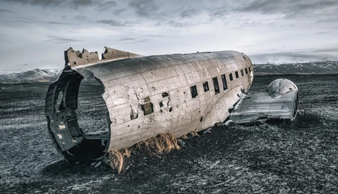 Abandoned aircraft fuselage lies on dark volcanic sand with mountains in the distance under overcast sky.