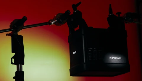 Studio lighting setup with a key light on stand and large flash unit mounted overhead against a red and yellow gradient backdrop.