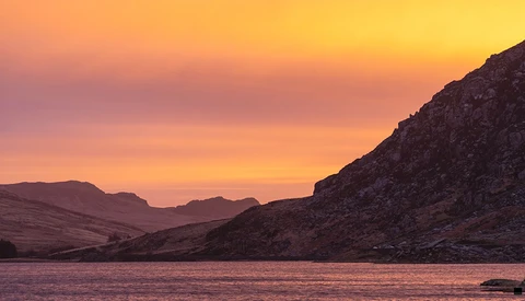 Rocky mountainous shoreline at sunset with warm golden and pink sky reflected in still water.