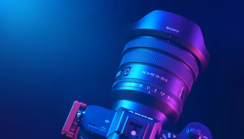 Sony camera lens mounted on camera body, illuminated with blue and magenta neon lighting against dark background.