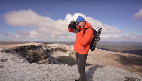 Photographer in orange jacket and blue cap holding camera, standing on white chalk cliff overlooking vast landscape.