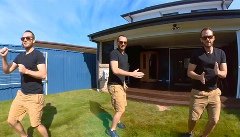 Three men jogging together on a sunny day in front of a modern house with a blue shipping container visible in the background.
