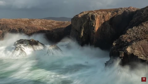 Turbulent ocean waves crash against rocky cliffs under overcast sky.