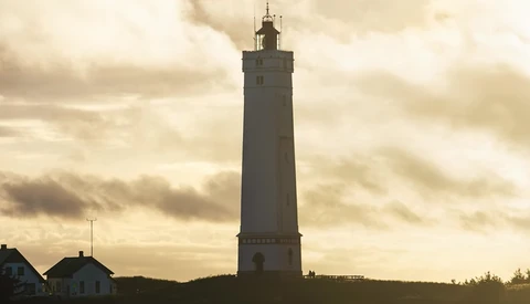 Tall cylindrical lighthouse with lantern room silhouetted against dramatic cloudy sky at dusk.