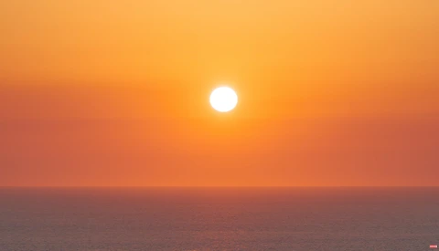 Bright sun descending toward the ocean horizon under a gradient orange and pink sky at sunset.