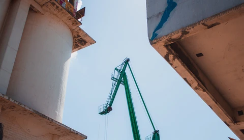 Green industrial crane extending upward against white cylindrical storage tanks and blue sky.