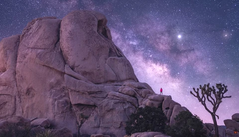 Lone figure standing on massive boulder beneath Milky Way galaxy at night.