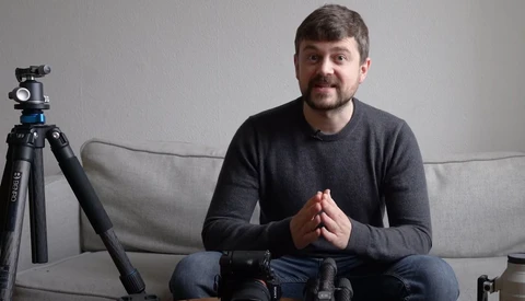 Man seated on a couch speaking to camera with tripod-mounted camera and lighting equipment visible in foreground.