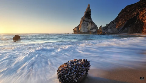 Dramatic coastal landscape with a towering rock formation rising from the ocean amid breaking waves at golden hour.
