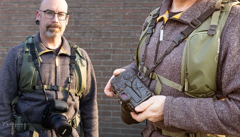 Two photographers wearing tactical vests and backpacks stand against a brick wall, one holding a camera and the other holding a gimbal stabilizer.