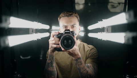 Photographer holding a DSLR camera directly toward the camera with symmetrical studio lighting in the background.