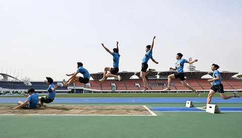 Young athletes in blue uniforms perform long jump attempts on an outdoor track and field stadium.
