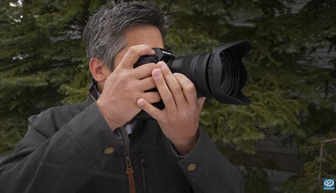 Photographer holding a DSLR camera with telephoto lens to his eye, wearing a dark jacket.