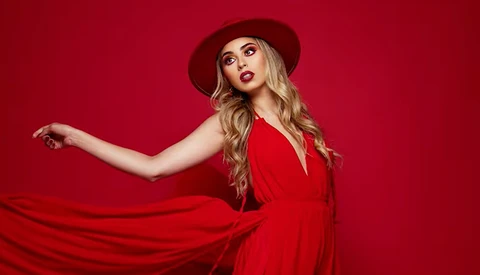 Woman in red dress and burgundy hat against solid red background, arm extended gracefully.