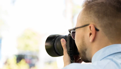 Photographer wearing glasses and light blue shirt holding DSLR camera to his face while shooting outdoors.