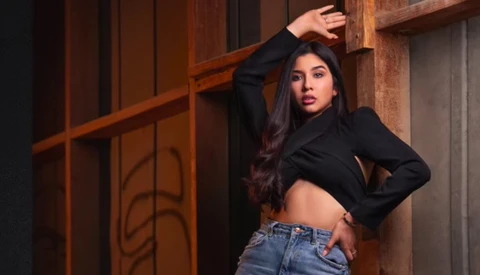 Woman in black crop top and jeans posed against wooden architectural elements with warm ambient lighting.