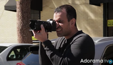 Photographer holding a DSLR camera with telephoto lens to his eye, composing a shot outdoors in bright sunlight.