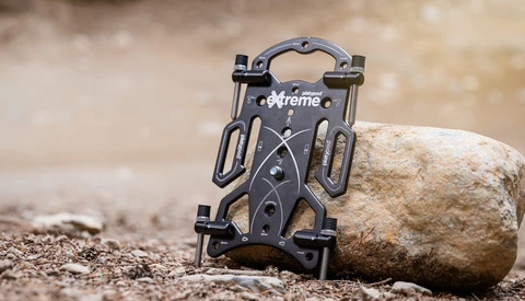 Black phone mount holder with grip arms positioned against a large stone on sandy ground.