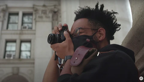 Photographer wearing glasses and a pink camera strap looks through a mirrorless camera viewfinder while positioned against a building facade.