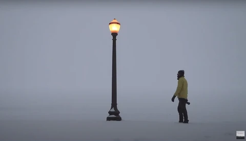 Person in yellow jacket standing near an illuminated street lamp against a gray sky.