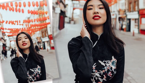 Portrait of a woman with long dark hair and red lipstick wearing a black jacket with embroidered dragons, shot against a street backdrop with orange lanterns.
