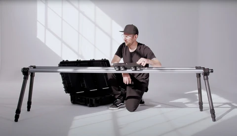Photographer crouching beside a large format camera mounted on a tripod in a bright studio with window light.