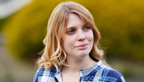 Young woman with shoulder-length blonde hair wearing a blue and white plaid shirt, looking toward the camera with a neutral expression.