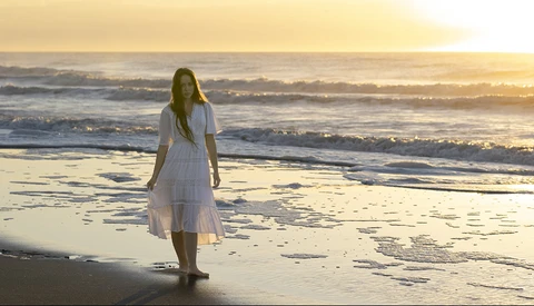 Woman in white dress standing in shallow ocean water during golden hour sunset.