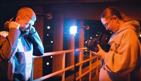 Two people on a metal railing at night, one singing into a microphone while the other operates a camera.