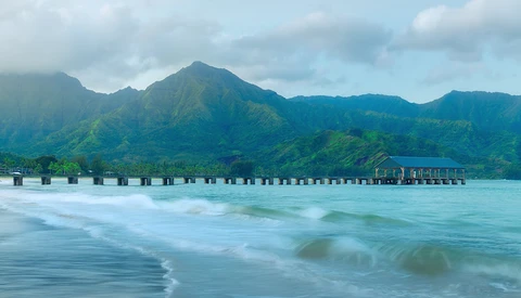 Wooden pier extending into turquoise ocean waters with green mountains rising dramatically behind it under partly cloudy skies.