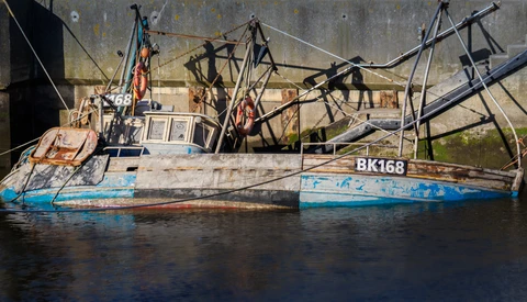 Weathered fishing vessel with registration number BK168 moored against a concrete seawall, its tangled rigging and nets silhouetted against gray water.