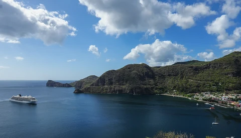 Tropical coastal bay with lush green mountainside, cruise ship anchored offshore, and small waterfront settlement nestled along the shoreline.