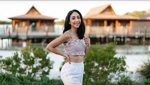 Woman posing in front of traditional wooden structures with water and vegetation in foreground.