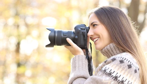 Female photographer holding a mirrorless camera with telephoto lens to her eye, smiling while shooting outdoors.