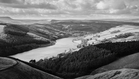 Monochrome landscape of a winding river valley surrounded by forested hills and open farmland under dramatic clouds.