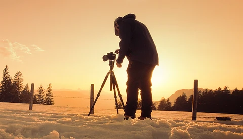 Silhouetted photographer operating a camera on tripod in snowy landscape at golden hour.