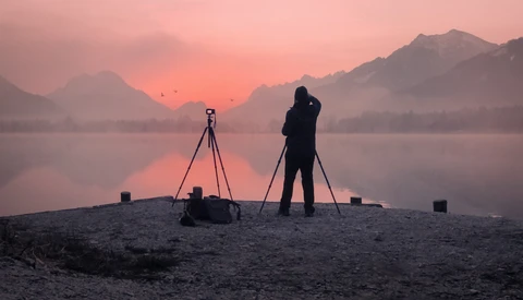 Photographer with tripod and camera silhouetted on beach at sunrise, facing misty mountains and still water.