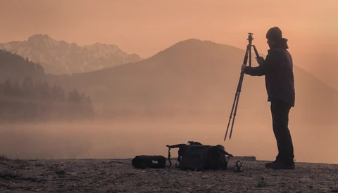 Silhouetted surveyor operating a theodolite on a tripod at sunrise, with misty mountains and forested terrain in the background.