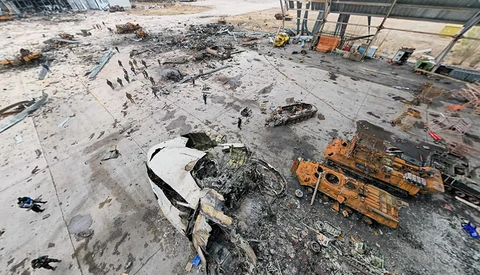 Aerial view of a destroyed white vehicle and heavy machinery on a debris-covered ground with scattered figures.