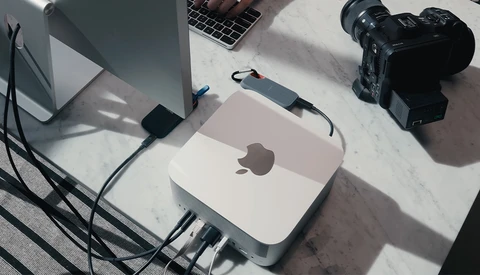 Overhead view of a creative workspace with Mac mini, monitor, laptop, video camera, and cables on marble surface.