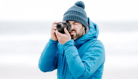 Bearded man in blue winter jacket and knit beanie holding vintage film camera to his face.