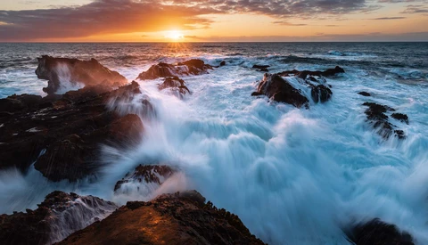 Waves crash against dark rocks at sunset, with soft motion blur creating ethereal white foam patterns.