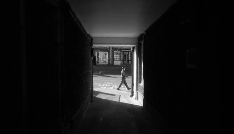 Silhouetted figure walking across a sunlit street, viewed through a dark doorway or corridor frame.