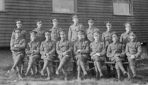 Fifteen uniformed soldiers arranged in three rows posing before a wooden building.