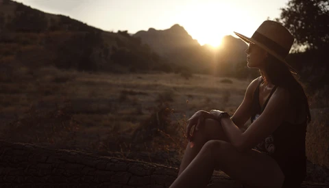 Woman wearing a hat sitting outdoors during golden hour sunset, gazing toward distant mountains.