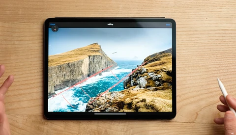 Tablet displaying a coastal landscape with dramatic cliffs and turquoise water, held by hands on a wooden surface.