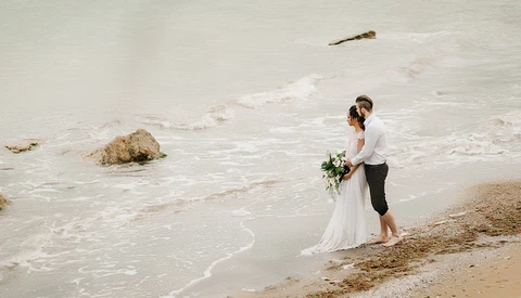 Bride and groom standing in shallow ocean waves on a beach, holding a bouquet.