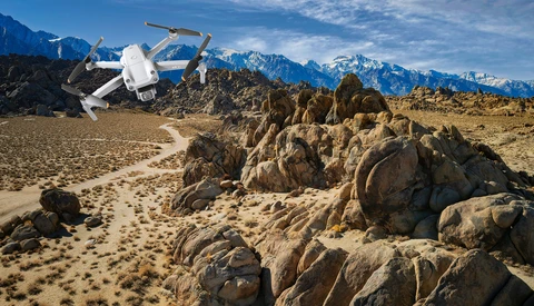 White drone hovering over rocky desert terrain with snow-capped mountains in the background.