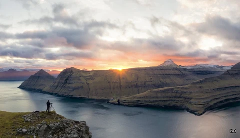Photographer standing on rocky outcrop overlooking fjord at sunset with snow-capped mountains.