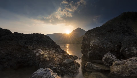 Rocky coastline framing a mountain and sun at golden hour with calm water between dark stone formations.